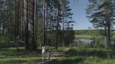 Anne ve küçük kızının hava manzarası ormanda ağaçların arasından nehre doğru bir patika boyunca yürüyor. Sahilde düğün kemeri olan bir rıhtım. Yaz güneşli bir gün