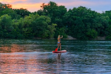 Gün batımında Dinyeper nehri boyunca kürek tahtasında (paddleboard, SUP) duran genç bir kadın.