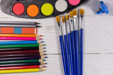 Beyaz tahta zemin üzerinde çizim araçları kümesi. Suluboya resimler, boya fırçaları, renkli kalemler, silgi ve masanda bileyici. Üst görünüm