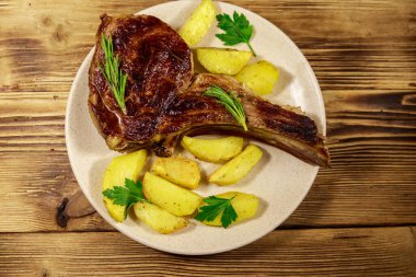 Kemiğin üstünde biftek, fırında patates ve ahşap masada otlar.