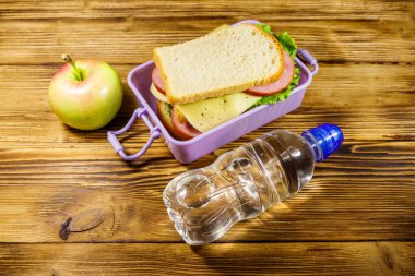 Sandviçli beslenme çantası, bir şişe su ve ahşap masada elma.
