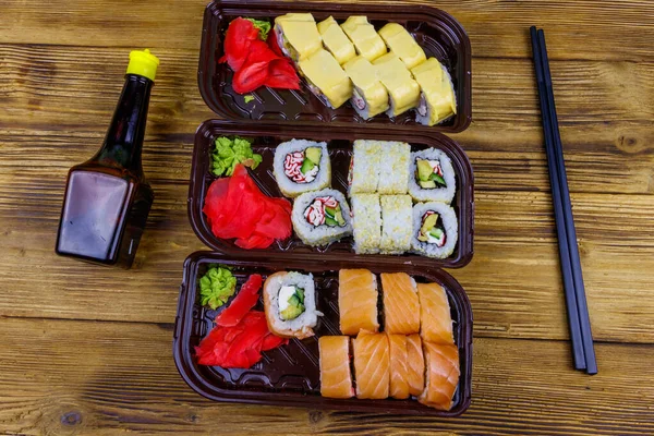 Set of sushi rolls in plastic boxes, soy sauce and chopsticks on wooden table. Top view. Sushi for take away or delivery of sushi in plastic containers