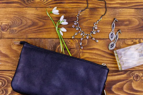 Women accessories on wooden background. Clutch bag, bottle of perfume, necklace, earrings and snowdrops on wood table. Beauty and fashion composition. Top view, flat lay