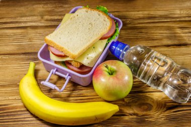 Sandviçli beslenme çantası, bir şişe su, muz ve elma ahşap bir masada.