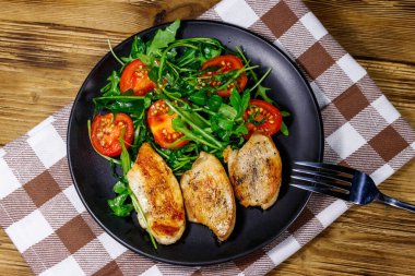 Kızarmış tavuk göğsü, taze roka salatası ve ahşap masada vişneli domates. Üst görünüm