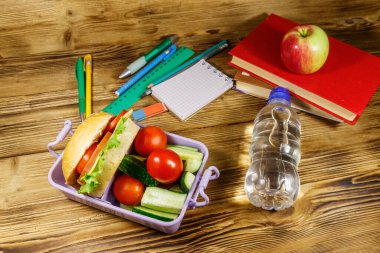 Okul konseptine geri dönelim. Okul malzemeleri, kitaplar, bir şişe su, elma ve hamburgerler ve taze sebzeler ahşap bir masada.