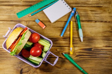 Okul konseptine geri dönelim. Okul malzemeleri, hamburgerler ve ahşap bir masada taze sebzelerle beslenme çantası. Üst görünüm