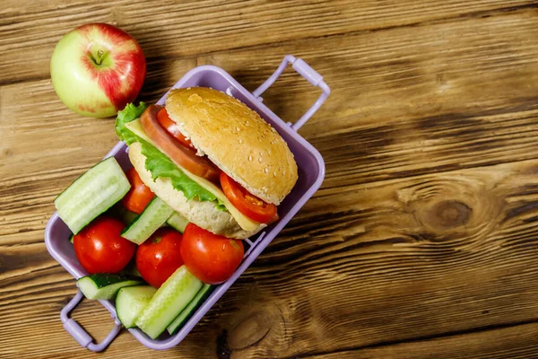 Elma, hamburger ve ahşap masada taze sebzeli beslenme çantası. Üst görünüm
