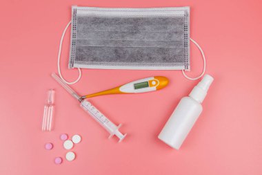 Koronavirüsü önleme ve tedavi etme kavramı. Tıbbi maske, el dezenfektanı, elektronik termometre, haplar, şırınga, pembe arka planda ampuller. Üst görünüm