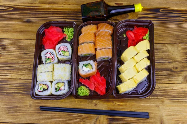 Set of sushi rolls in plastic boxes, soy sauce and chopsticks on wooden table. Top view. Sushi for take away or delivery of sushi in plastic containers