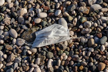 Deniz kıyısındaki çakıl taşı plajında kullanılmış tek kullanımlık tıbbi maske. COVID-19 sırasında israf. Koronavirüs salgınından kaynaklanan kirlilik
