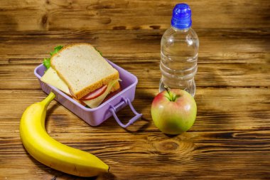 Sandviçli beslenme çantası, bir şişe su, muz ve elma ahşap bir masada.