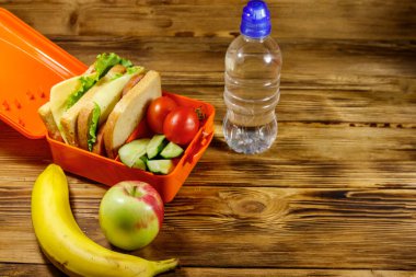 Bir şişe su, elma, muz ve sandviç ve taze sebzelerle birlikte ahşap bir masada.