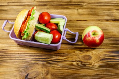 Elma, beslenme çantası, hamburgerler ve ahşap bir masada taze sebzeler.