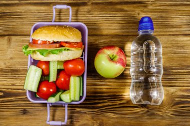 Bir şişe su, elma ve beslenme çantası, içinde hamburgerler ve ahşap bir masada taze sebzeler. Üst görünüm