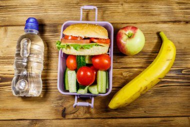Bir şişe su, elma, muz ve hamburgerli beslenme çantası ve tahta bir masada taze sebzeler. Üst görünüm