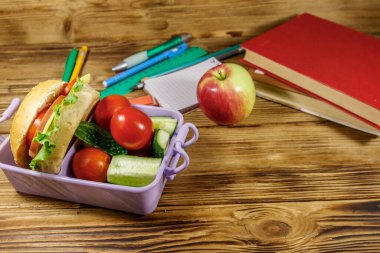Okul konseptine geri dönelim. Okul gereçleri, kitaplar, elma ve beslenme çantası. Hamburgerler ve taze sebzeler ahşap bir masada.