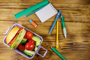 Okul konseptine geri dönelim. Okul malzemeleri, hamburgerler ve ahşap bir masada taze sebzelerle beslenme çantası. Üst görünüm