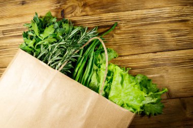 Yeşil soğanlı kağıt torbası, biberiye, marul yaprakları ve ahşap masada maydanoz. Üst Manzara. Sağlıklı gıda ve market alışverişi kavramı