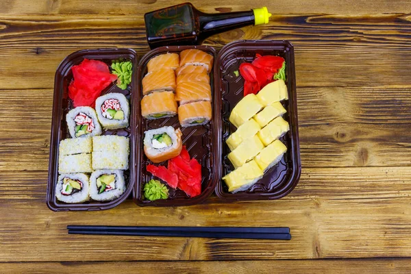 Set of sushi rolls in plastic boxes, soy sauce and chopsticks on wooden table. Top view. Sushi for take away or delivery of sushi in plastic containers