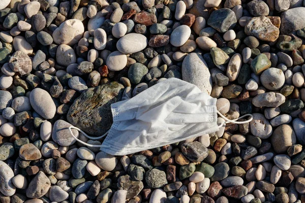 Dirty used disposable medical face mask on a pebble beach by sea. Waste during COVID-19. Pollution due coronavirus pandemic