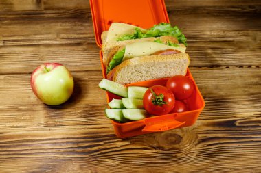 Elma ve beslenme çantasıyla sandviç ve taze sebzeler ahşap bir masada.
