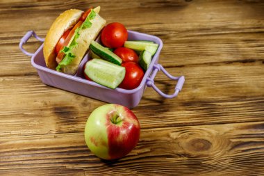 Elma, beslenme çantası, hamburgerler ve ahşap bir masada taze sebzeler.