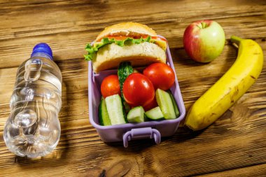 Bir şişe su, elma, muz ve hamburgerli beslenme çantası ve ahşap bir masada taze sebze.