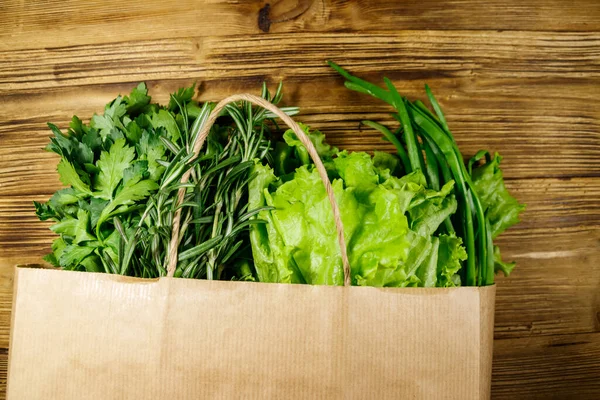 Paper bag with green onion, rosemary, lettuce leaves and parsley on wooden table. Top view. Healthy food and grocery shopping concept
