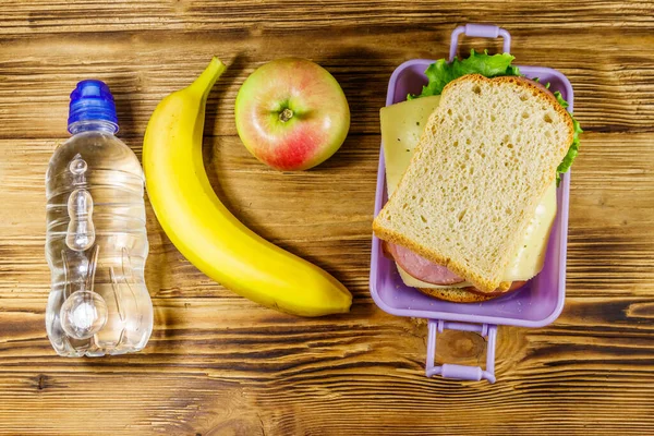 Sandviçli beslenme çantası, bir şişe su, muz ve elma ahşap bir masada. Üst görünüm
