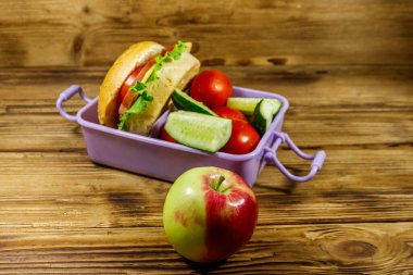 Elma, beslenme çantası, hamburgerler ve ahşap bir masada taze sebzeler.