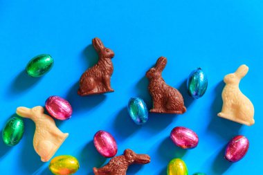 Folyo içinde Paskalya çikolatası yumurtaları ve mavi arka planda Paskalya çikolatası tavşanları. Üst görünüm