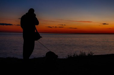 Gün batımında nehir kıyısında fotoğraf makinesi ve köpeği olan bir adamın silueti.