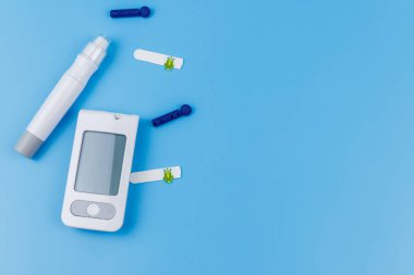 Dijital glukometre, tükenmez kalem, tek kullanımlık iğneler ve pastel mavi arka planda test şeritleri. Üst görünüm, boşluğu kopyala. Diyabet kavramı