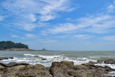 Mavi gökyüzünün altında, dalgalar sahildeki kayaları dövüyor.