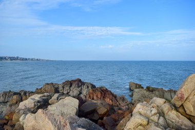 Mavi gökyüzünün altında, dalgalar sahildeki kayaları dövüyor.