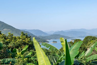 Mavi gökyüzünün altında, göl dağlar ve ormanlarla çevrilidir.