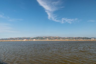 Mavi gökyüzünün altındaki kıyı şeridi, denizin karşısında dağlar ve rüzgar enerjisi var.