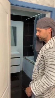 Sakallı adam banyoya giriyor, bornozunu çıkarıyor ve kaslı sırtını gösteriyor. Kafasına bir havlu sarılmış ve pijama pantolon giyiyor. Sahnede özbakım, ev konforu ve rahatlama vurgulanıyor..