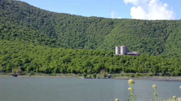Un petit château dans les collines verdoyantes sur les rives du lac. Ciel bleu, nuages, balançant dans l'herbe du premier plan — Video