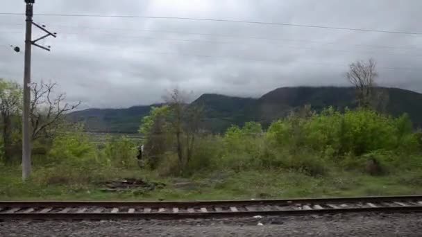 Utsikten från fönstret på tåget på rälsen och de grönskande bergen i fjärran på en bakgrund av vita moln. Kaukasus, Georgien. — Stockvideo