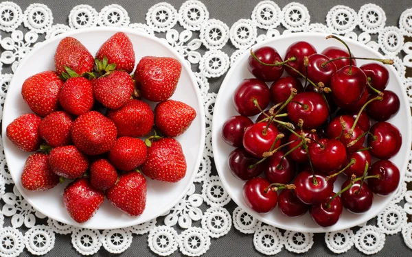Plate with strawberries and a plate of red cherries on a lacy napkin, top view — Stock Photo, Image