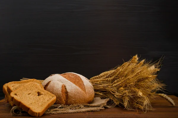Деревянный стол с круглым и квадратным ржаным хлебом, сноп, черный фон, с местом для текста — стоковое фото