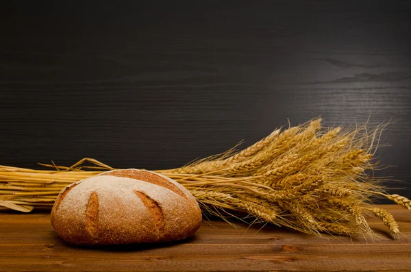 Круглый ржаной хлеб и сноп на деревянном столе, черный фон, место для текста — стоковое фото