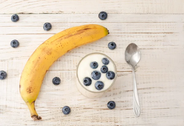 Plátano maduro, yogur con arándanos y cuchara, mesa ligera de madera — Foto de Stock