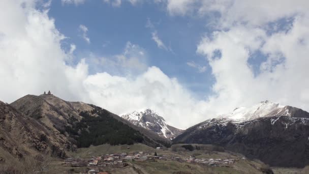 Weergave van het bergdorp en de tempel van de Heilige Drievuldigheid-Gergeti in de bergen op een achtergrond van witte wolken drijven — Stockvideo