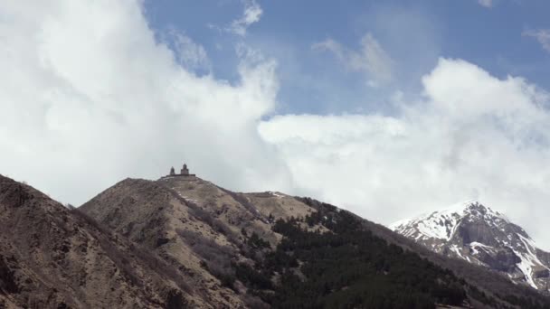 Blick auf die Berge und den antiken Tempel der heiligen Dreifaltigkeit Gergeti auf einem Hintergrund aus weißen Wolken schweben. Georgien, Kaukasus. — Stockvideo