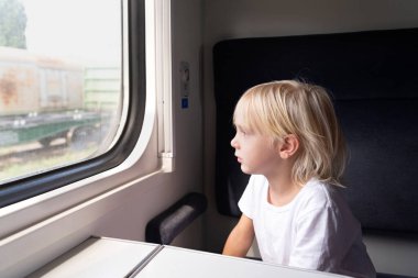 Küçük sarışın çocuk düşünceli bir şekilde tren penceresinden dışarı bakıyor. Çocuklarla tren yolculuğu..