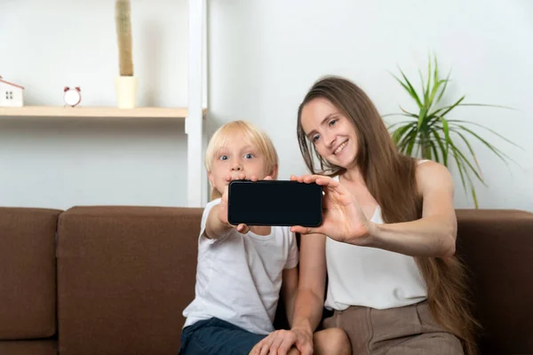 Mom and son take selfie while sitting on couch at home. Mom and child showing a phone screen