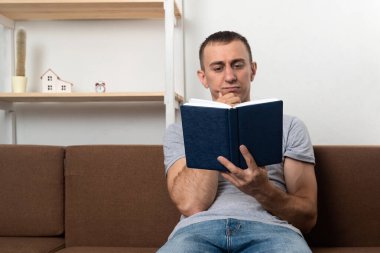 Genç adam oturma odasında kanepede otururken düşünceli bir şekilde kitap okuyor.